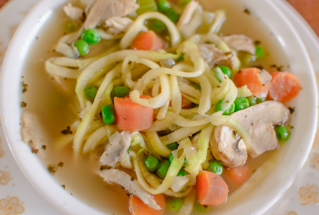 A close up of a bowl of chicken soup made with zucchini noodles, chicken and other colorful vegetables.